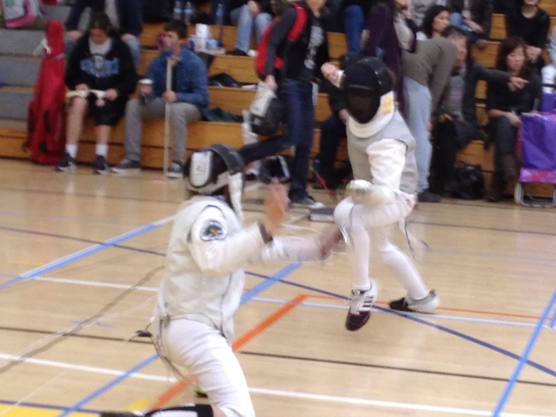 Marin Fencing Academy — A masked fencer attacking on a gym floor with spectators watching from the bleachers