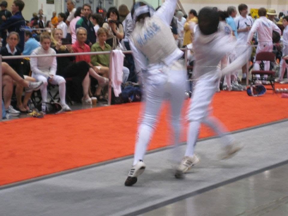 Marin Fencing Academy — Two fencers mid-exchange on a red piste at a tournament, spectators behind