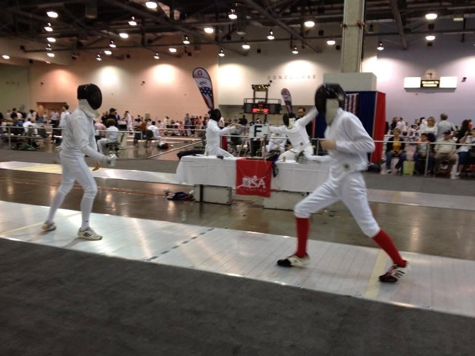Marin Fencing Academy — Two masked fencers advancing on a strip at a national tournament, referee and scoring box behind