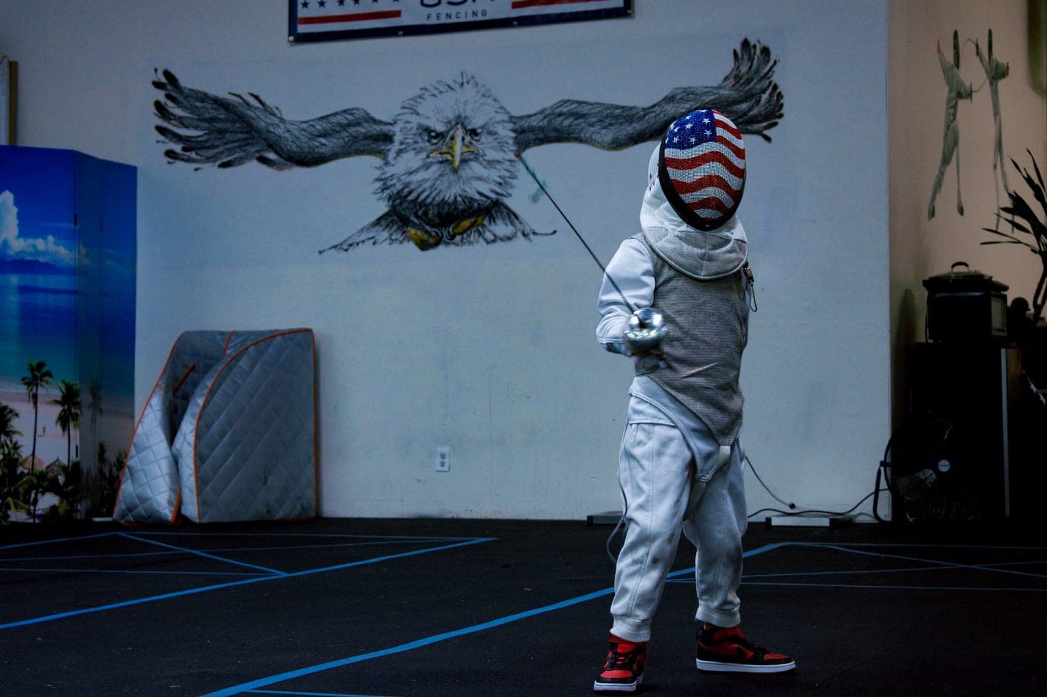 A masked fencer on the piste at Marin Fencing Academy, beneath the club’s painted eagle